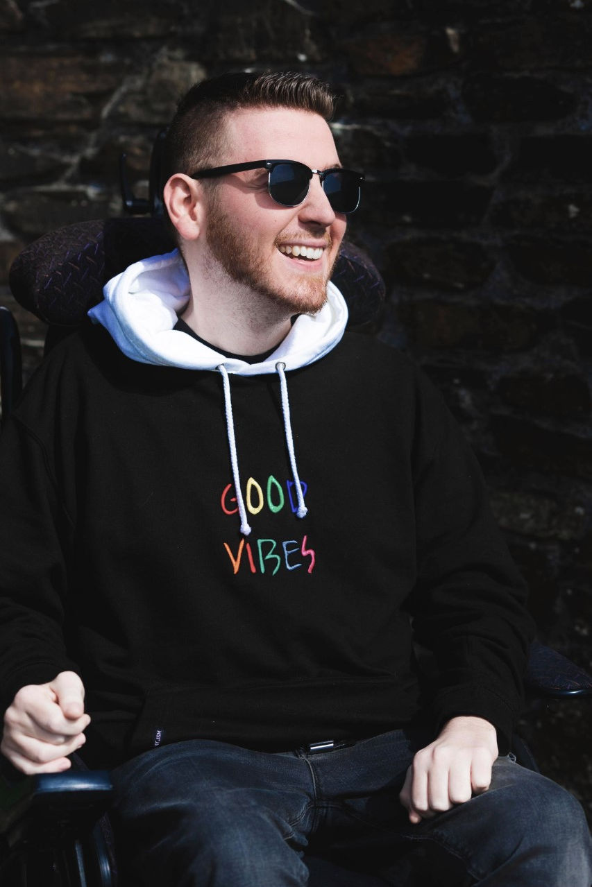 Professional image taken of Ross smiling, wearing sunglasses and a black hoody that says "good vibes"