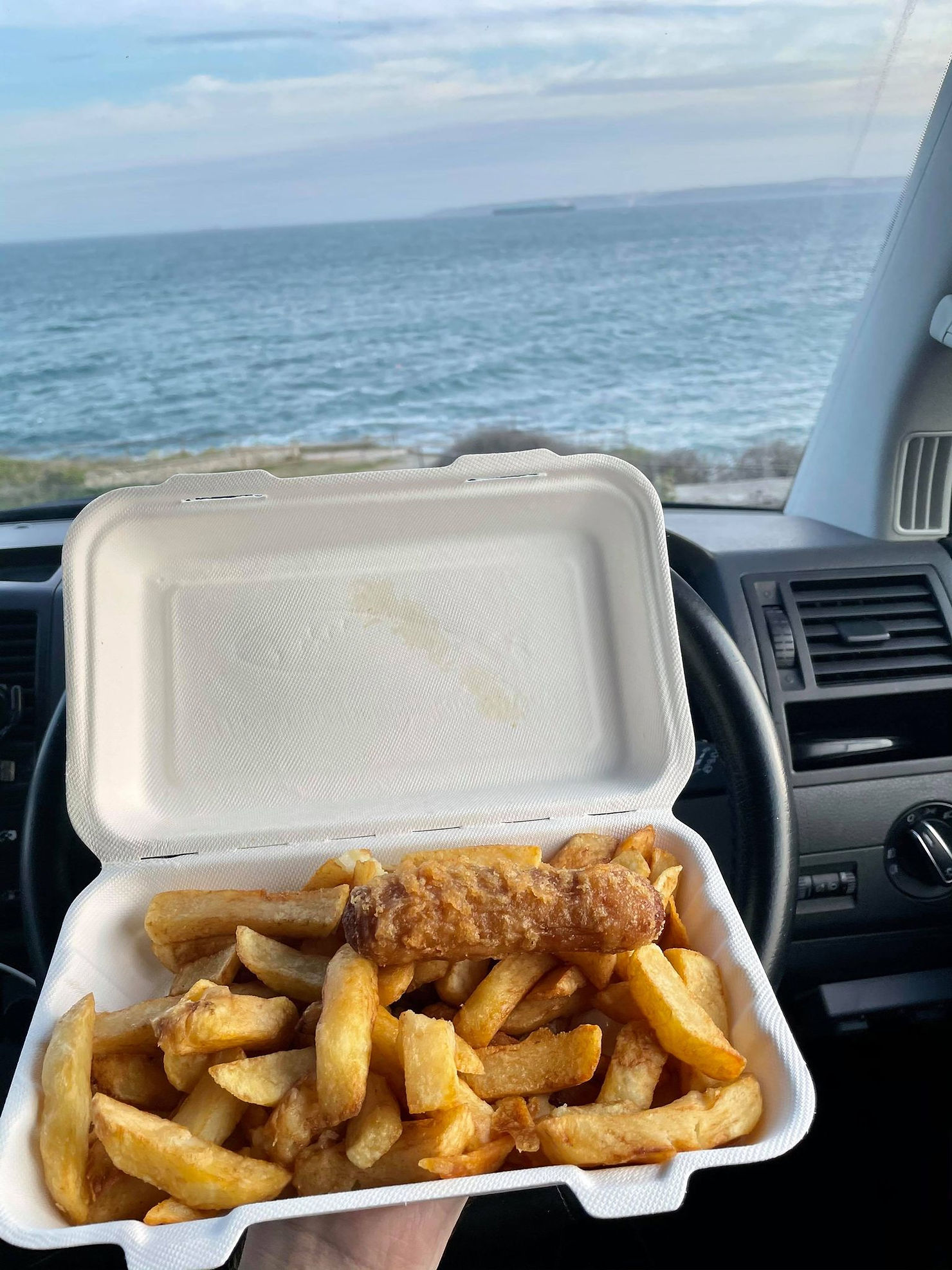 View from the drivers seat, Ross's wrist holding a tray of sausage and chips with the sea in the background