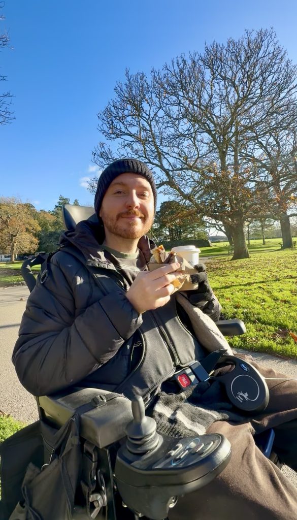 Ross sat in his wheelchair in the park, holding a hot drink and cake. Wrapped up in winter hat and coat.