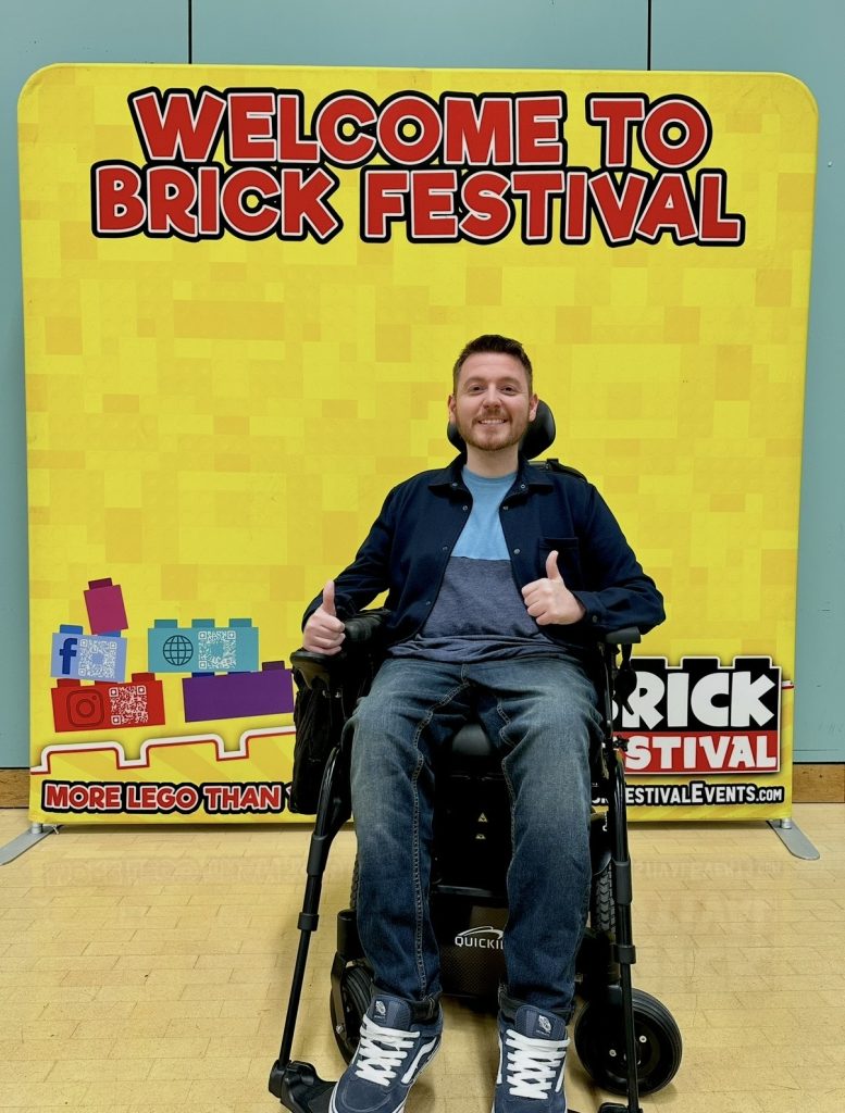 Ross sat in his wheelchair in front of a large yellow sign that says "welcome to brick festival"