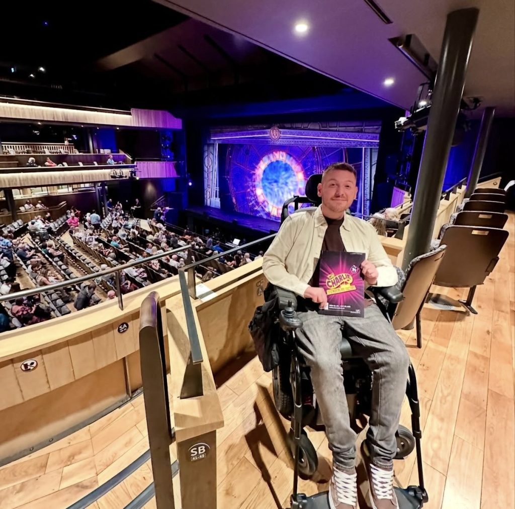 Ross inside the Hall For Cornwall theatre, holding a Charlie & The Chocolate Factory programme, with the stage in the background.